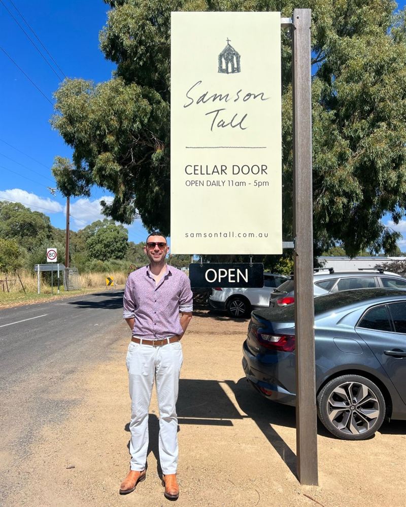 a man stands in front of a cellar door sign in a treelined carpark